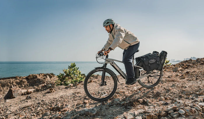 A rider takes a Fiido Nomads touring e-bike equipped with rear panniers across a rocky coastal trail.